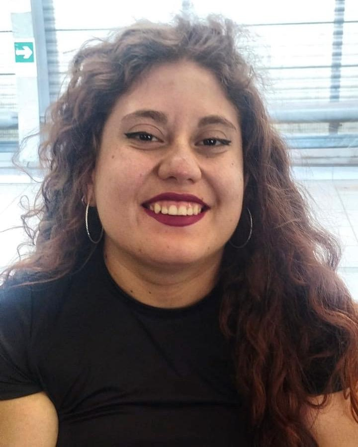 Persona sonriendo con cabello rizado y una camiseta oscura, en un entorno interior con luz natural.