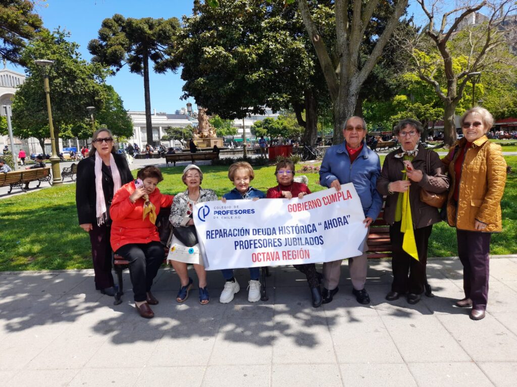 Un grupo de profesores jubilados de la Octava Región de Chile se manifiestan en un parque, sosteniendo un cartel que dice: 'Gobierno cumpla. Reparación deuda histórica "ahora". Profesores jubilados Octava Región.' Están sentados y de pie, mostrando su apoyo a la causa por la reparación de la deuda histórica de los docentes.