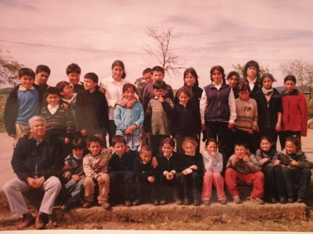 Fotografía de grupo escolar al aire libre, con un docente sentado al frente y rodeado de niños y niñas. La imagen muestra una escena típica de un colegio rural, donde los estudiantes posan juntos, vestidos de manera informal y en un entorno natural.